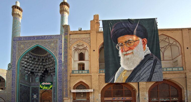 Detalle de la fachada y entrada de la Gran Mezquita de Isfahán (Irán), con su característico arco decorado con azulejos azules y minaretes ornamentados. A la derecha, un gran retrato de Alí Jamenei, líder supremo del país, cuelga sobre un edificio de ladrillo con arcos y motivos geométricos