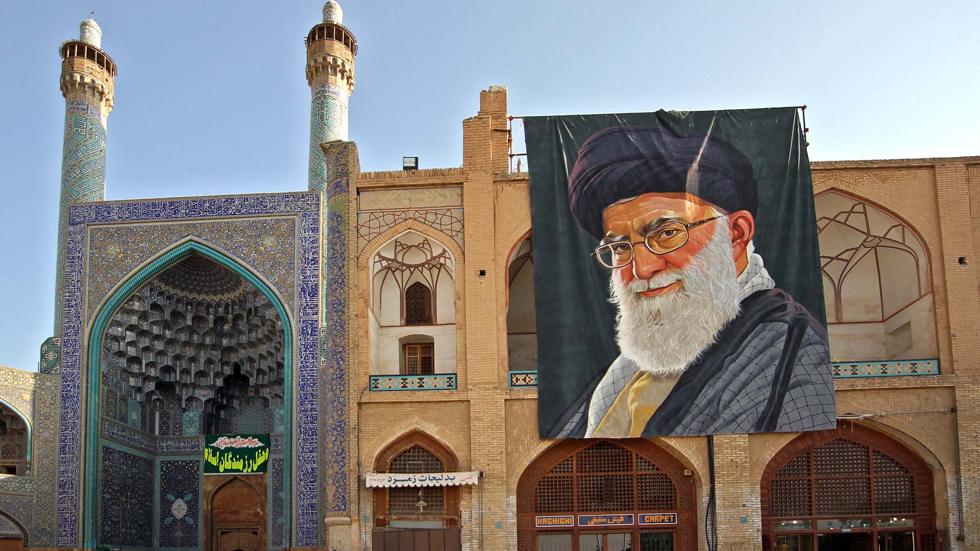 Detalle de la fachada y entrada de la Gran Mezquita de Isfahán (Irán), con su característico arco decorado con azulejos azules y minaretes ornamentados. A la derecha, un gran retrato de Alí Jamenei, líder supremo del país, cuelga sobre un edificio de ladrillo con arcos y motivos geométricos