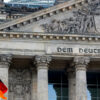 German flag (with horizontal stripes in black, red, and yellow), waving in front of the Reichstag building in Berlin during a sunset, with the sky painted in warm tones and clouds. Germany