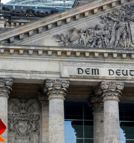 Bandera de Alemania (con franjas horizontales en negro, rojo y amarillo), ondeando frente al edificio del Reichstag en Berlín durante un día nublado. De fondo, se observa la cúpula de cristal del edificio. Alemania