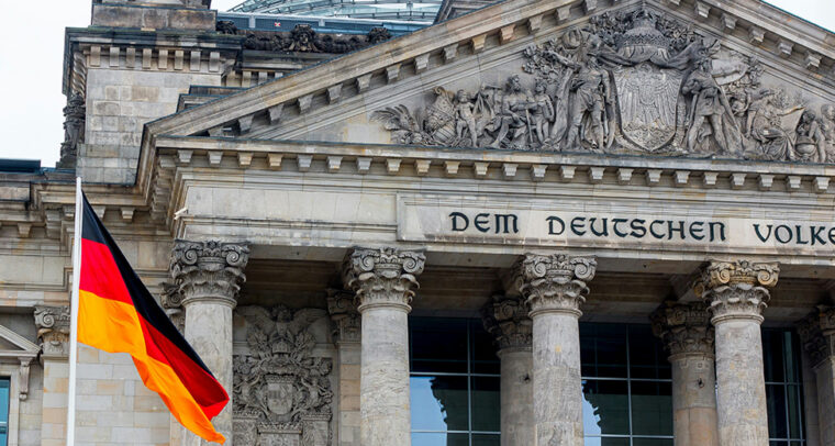 Bandera de Alemania (con franjas horizontales en negro, rojo y amarillo), ondeando frente al edificio del Reichstag en Berlín durante un día nublado. De fondo, se observa la cúpula de cristal del edificio. Alemania