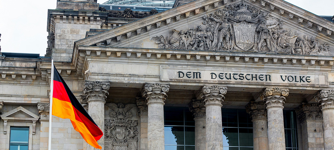 German flag (with horizontal stripes in black, red, and yellow), waving in front of the Reichstag building in Berlin during a sunset, with the sky painted in warm tones and clouds. Germany