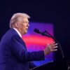 Donald Trump (de lado con un traje azulado, frente a un atril con micrófono y con el fondo de una pantalla borrosa con colores rojizos y púrpuras) se dirige a los asistentes del AmericaFest 2024 en el Centro de Convenciones de Phoenix (Arizona, EEUU), el 22 de diciembre de 2024. Foto: Gage Skidmore (CC BY-SA 2.0)