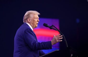 Donald Trump (de lado con un traje azulado, frente a un atril con micrófono y con el fondo de una pantalla borrosa con colores rojizos y púrpuras) se dirige a los asistentes del AmericaFest 2024 en el Centro de Convenciones de Phoenix (Arizona, EEUU), el 22 de diciembre de 2024. Foto: Gage Skidmore (CC BY-SA 2.0)