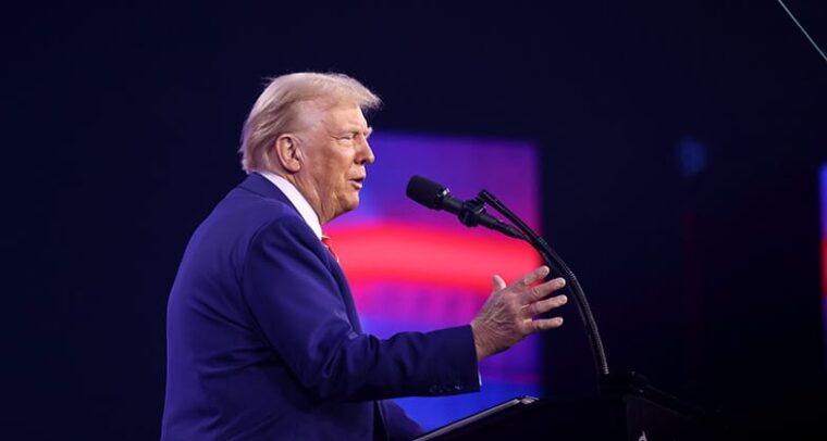 Donald Trump (de lado con un traje azulado, frente a un atril con micrófono y con el fondo de una pantalla borrosa con colores rojizos y púrpuras) se dirige a los asistentes del AmericaFest 2024 en el Centro de Convenciones de Phoenix (Arizona, EEUU), el 22 de diciembre de 2024. Foto: Gage Skidmore (CC BY-SA 2.0)