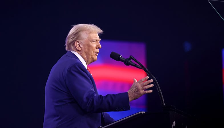 Donald Trump (de lado con un traje azulado, frente a un atril con micrófono y con el fondo de una pantalla borrosa con colores rojizos y púrpuras) se dirige a los asistentes del AmericaFest 2024 en el Centro de Convenciones de Phoenix (Arizona, EEUU), el 22 de diciembre de 2024. Foto: Gage Skidmore (CC BY-SA 2.0)