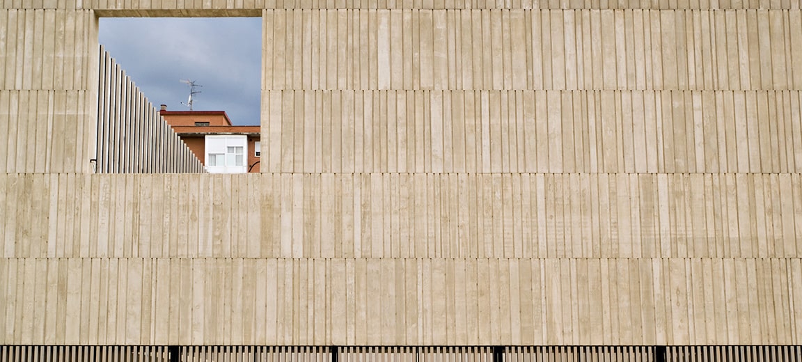 Fachada de un edificio de hormigón gris con textura vertical y una gran abertura cuadrada que enmarca un edificio de fondo con techos rojos. En la parte inferior, se observa una reja de lamas verticales de color negro. Yihadista