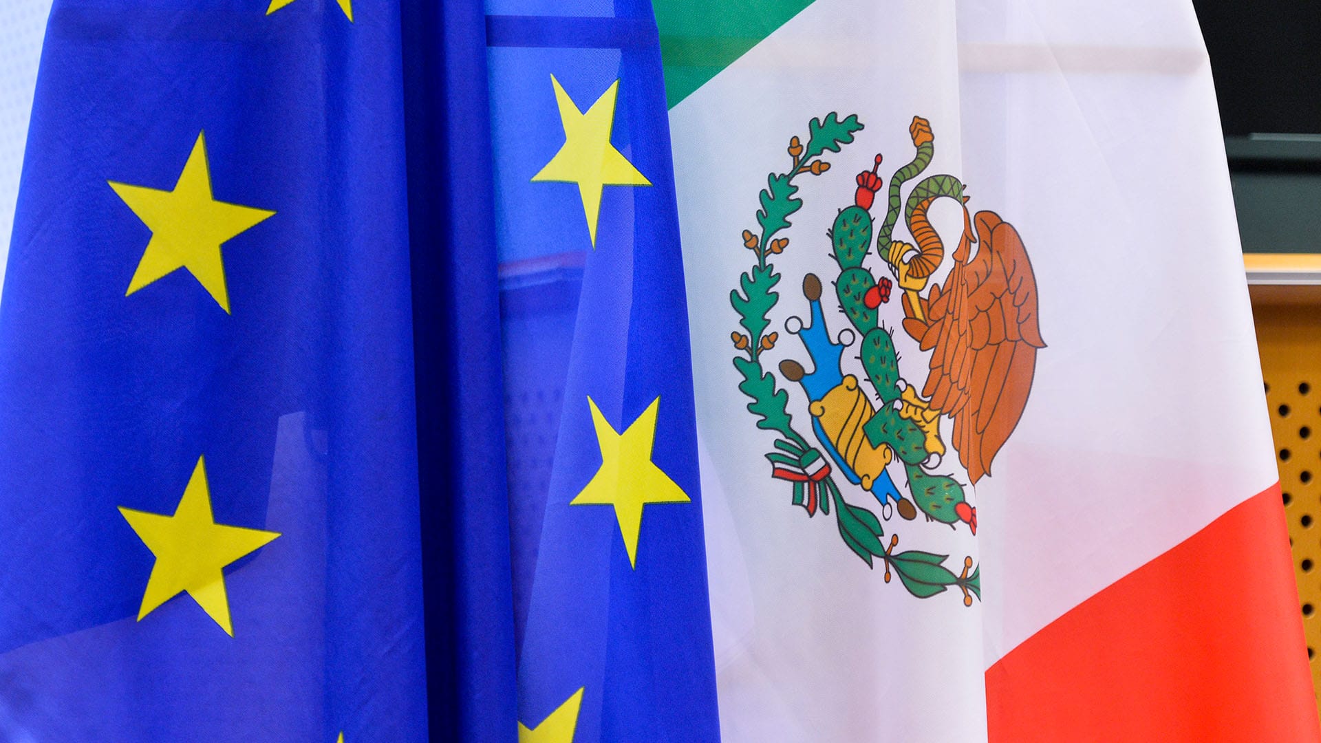 Flags of the European Union (blue with a circle of yellow stars) and Mexico (tricolour with green, white, and red stripes, featuring a coat of arms with an eagle and a serpent) at the 25th Meeting of the Mexico-European Union Joint Parliamentary Committee in 2018. Mexico