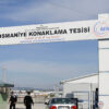 Security checkpoint at the entrance of the Osmaniye Accommodation Facility in Turkey. A large sign overhead shows the facility’s name in three languages (English, Turkish and Arabic), along with the Turkish flag and AFAD (Turkey’s Disaster and Emergency Management Authority) logo. A silver car is stopped at the gate as two security officers check the area. A man stands near a white building, with blue fences and barriers around. Refugees