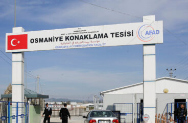 Security checkpoint at the entrance of the Osmaniye Accommodation Facility in Turkey. A large sign overhead shows the facility’s name in three languages (English, Turkish and Arabic), along with the Turkish flag and AFAD (Turkey’s Disaster and Emergency Management Authority) logo. A silver car is stopped at the gate as two security officers check the area. A man stands near a white building, with blue fences and barriers around. Refugees