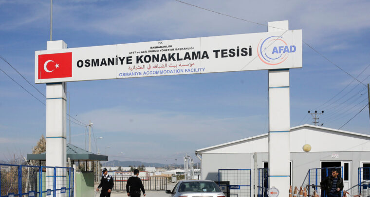 Security checkpoint at the entrance of the Osmaniye Accommodation Facility in Turkey. A large sign overhead shows the facility’s name in three languages (English, Turkish and Arabic), along with the Turkish flag and AFAD (Turkey’s Disaster and Emergency Management Authority) logo. A silver car is stopped at the gate as two security officers check the area. A man stands near a white building, with blue fences and barriers around. Refugees