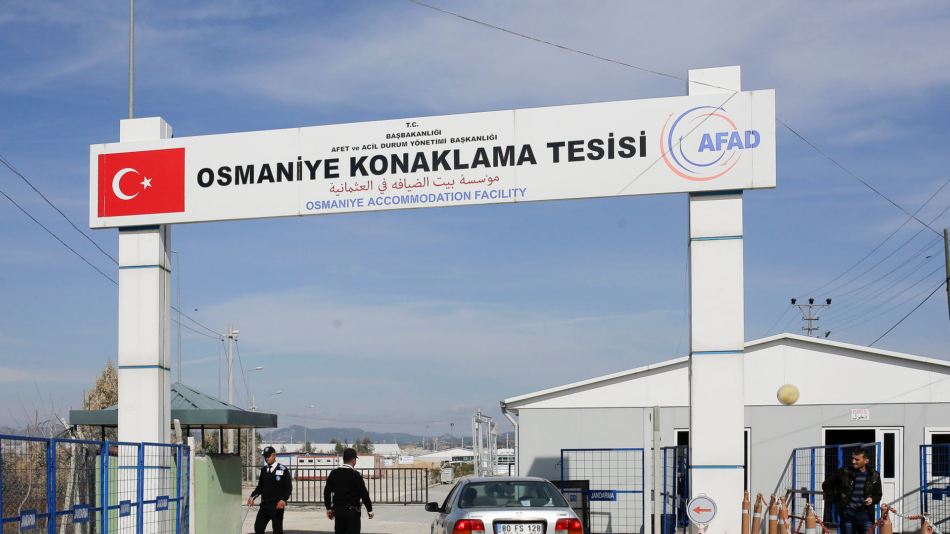 Security checkpoint at the entrance of the Osmaniye Accommodation Facility in Turkey. A large sign overhead shows the facility’s name in three languages (English, Turkish and Arabic), along with the Turkish flag and AFAD (Turkey’s Disaster and Emergency Management Authority) logo. A silver car is stopped at the gate as two security officers check the area. A man stands near a white building, with blue fences and barriers around. Refugees