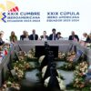 Jefes de Estado de países iberoamericanos participan en la XXIX Cumbre Iberoamericana en Ecuador 2023-2024, en la ciudad de Cuenca. Se observa una mesa en forma de U, decorada con flores y banderas nacionales. En el centro, los mandatarios principales presiden la sesión frente a un fondo con el logo oficial del evento, rodeado por ambos lados por las banderas de los países asistentes, mientras los demás líderes y delegados escuchan, toman notas y utilizan dispositivos electrónicos.