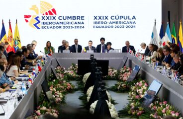 Jefes de Estado de países iberoamericanos participan en la XXIX Cumbre Iberoamericana en Ecuador 2023-2024, en la ciudad de Cuenca. Se observa una mesa en forma de U, decorada con flores y banderas nacionales. En el centro, los mandatarios principales presiden la sesión frente a un fondo con el logo oficial del evento, rodeado por ambos lados por las banderas de los países asistentes, mientras los demás líderes y delegados escuchan, toman notas y utilizan dispositivos electrónicos.