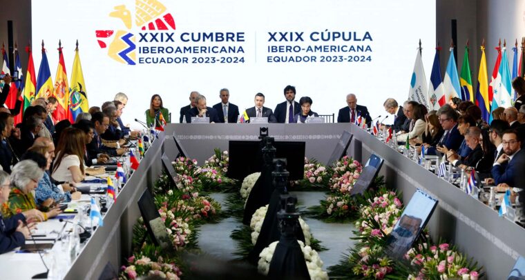 Jefes de Estado de países iberoamericanos participan en la XXIX Cumbre Iberoamericana en Ecuador 2023-2024, en la ciudad de Cuenca. Se observa una mesa en forma de U, decorada con flores y banderas nacionales. En el centro, los mandatarios principales presiden la sesión frente a un fondo con el logo oficial del evento, rodeado por ambos lados por las banderas de los países asistentes, mientras los demás líderes y delegados escuchan, toman notas y utilizan dispositivos electrónicos.