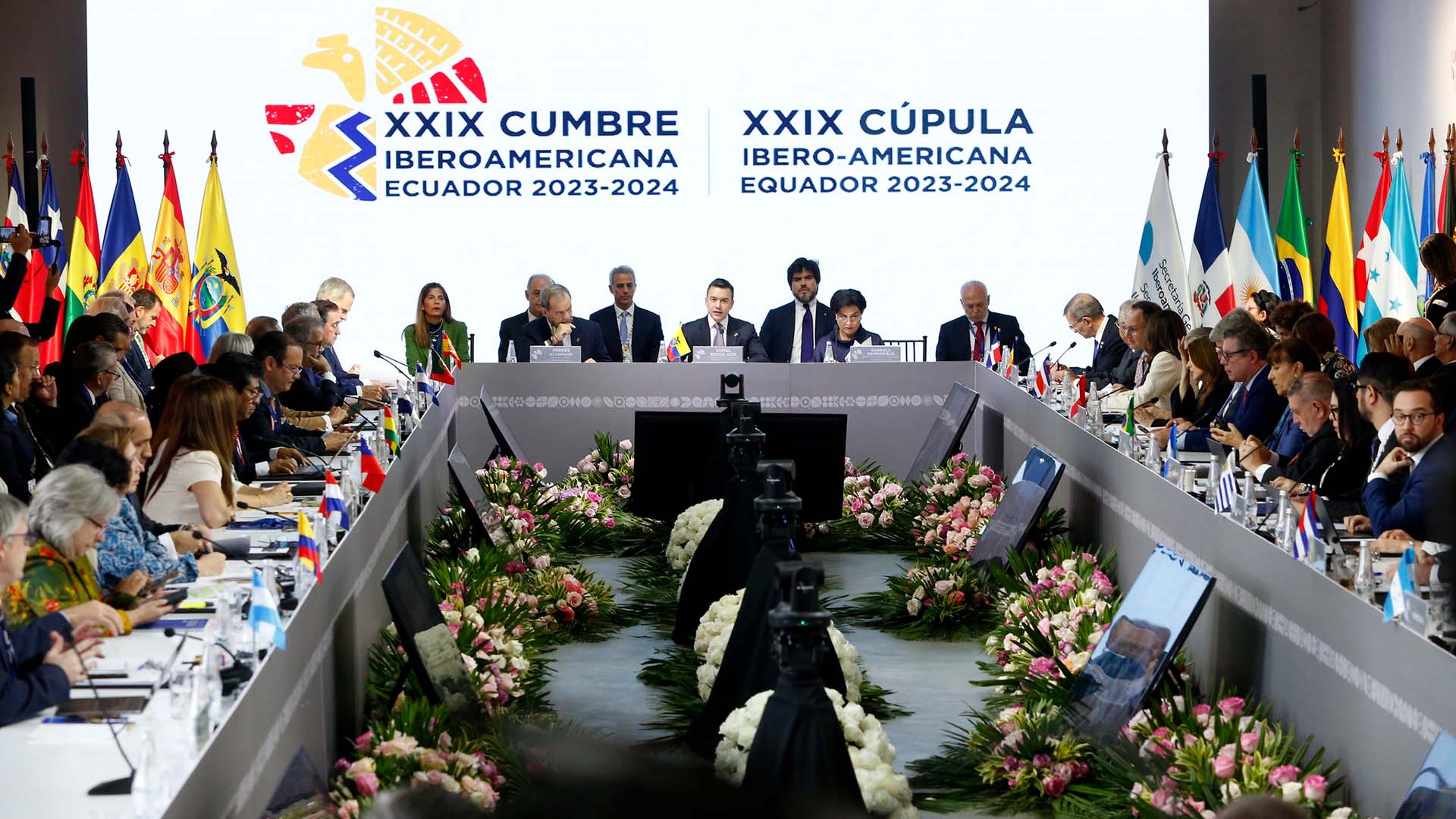 Jefes de Estado de países iberoamericanos participan en la XXIX Cumbre Iberoamericana en Ecuador 2023-2024, en la ciudad de Cuenca. Se observa una mesa en forma de U, decorada con flores y banderas nacionales. En el centro, los mandatarios principales presiden la sesión frente a un fondo con el logo oficial del evento, rodeado por ambos lados por las banderas de los países asistentes, mientras los demás líderes y delegados escuchan, toman notas y utilizan dispositivos electrónicos.