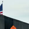 A section of the border wall between the United States and Mexico, located in Texas. In the foreground, the dark grey metal wall is visible, along with an orange warning sign that reads "Road closed 1000 ft." Behind the wall, a crane partially displays a United States flag against a cloudy sky. Deportation