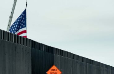 A section of the border wall between the United States and Mexico, located in Texas. In the foreground, the dark grey metal wall is visible, along with an orange warning sign that reads "Road closed 1000 ft." Behind the wall, a crane partially displays a United States flag against a cloudy sky. Deportation