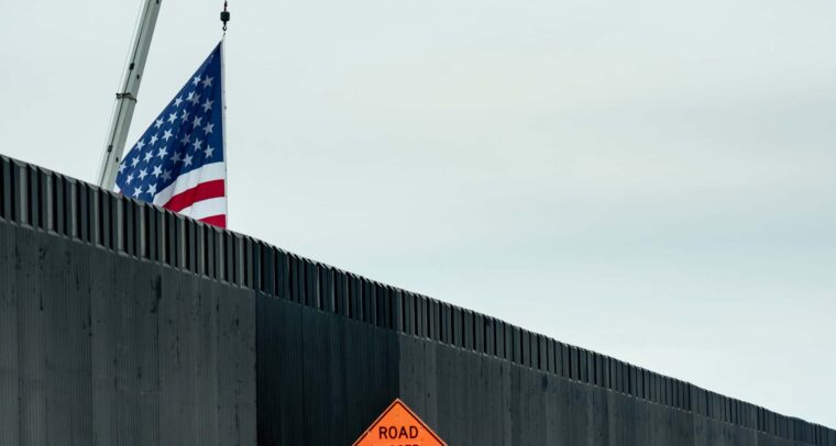 Tramo del muro fronterizo entre Estados Unidos y México, situado en Texas. En primer plano, se observa el muro de metal gris oscuro y una señal de advertencia naranja que indica "Road closed 1000 ft" ("Carretera cerrada en 1000 pies"). Detrás del muro, una grúa sostiene, parcialmente visible, una bandera de Estados Unidos contra un cielo nublado. Deportación