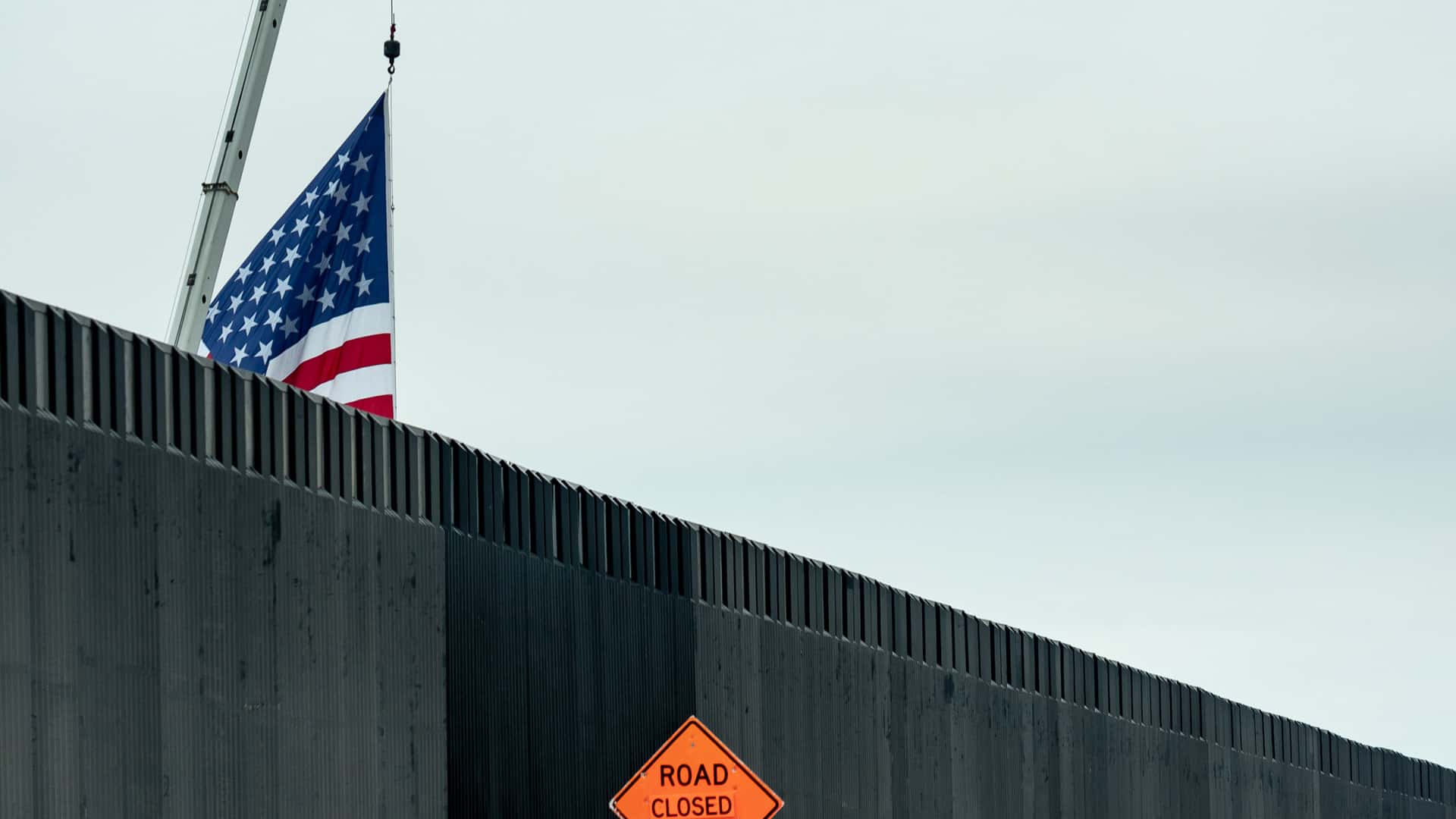 A section of the border wall between the United States and Mexico, located in Texas. In the foreground, the dark grey metal wall is visible, along with an orange warning sign that reads "Road closed 1000 ft." Behind the wall, a crane partially displays a United States flag against a cloudy sky. Deportation