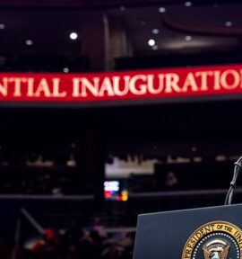 Donald Trump, presidente de los Estados Unidos, está de pie frente a un podio con el sello presidencial. Está vestido con un traje azul, una camisa blanca y una corbata de tono burdeos. De fondo, se aprecian las gradas del estadio Capitol One Arena en Washington (EE. UU.) y dos bandas de luz rojas con estrellas blancas y la frase "60th Presidential Inauguration" (60ª Inauguración Presidencial). Trump