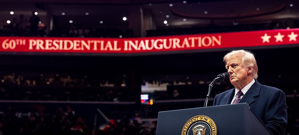 Donald Trump, presidente de los Estados Unidos, está de pie frente a un podio con el sello presidencial. Está vestido con un traje azul, una camisa blanca y una corbata de tono burdeos. De fondo, se aprecian las gradas del estadio Capitol One Arena en Washington (EE. UU.) y dos bandas de luz rojas con estrellas blancas y la frase "60th Presidential Inauguration" (60ª Inauguración Presidencial). Trump