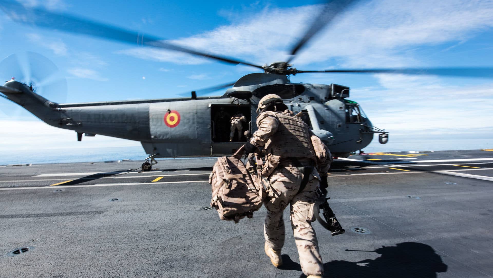 Primer plano de un soldado vestido con el uniforme militar español, con una mochila en la mano izquierda, corriendo hacia un helicóptero de la Armada Española durante operaciones anfibias desde el barco de cuatro anclas FS Mistral, como parte del ejercicio Brilliant Mariner 17, en un día soleado. Gasto de defensa