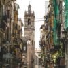 Valencia city street during the day. In the foreground, at the bottom, people are walking in different directions. Shops can be seen on either side. In the background, a church tower stands out against the skyline; it is built of stone, with a bell and a pointed top. Middle class