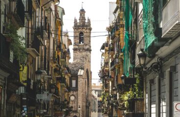 Valencia city street during the day. In the foreground, at the bottom, people are walking in different directions. Shops can be seen on either side. In the background, a church tower stands out against the skyline; it is built of stone, with a bell and a pointed top. Middle class