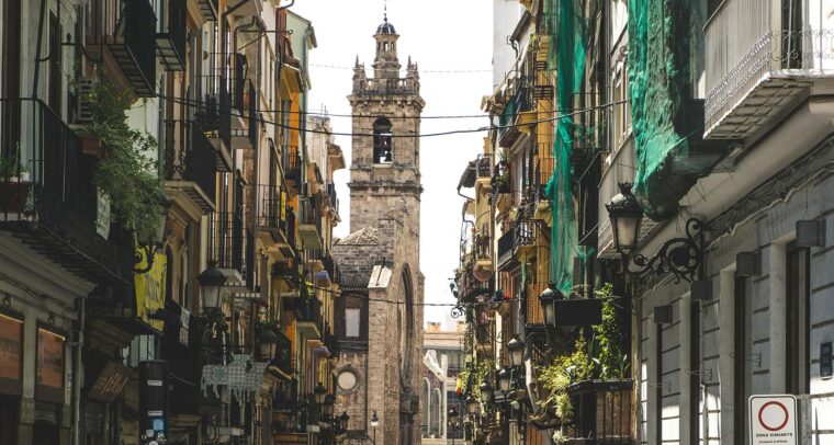 Valencia city street during the day. In the foreground, at the bottom, people are walking in different directions. Shops can be seen on either side. In the background, a church tower stands out against the skyline; it is built of stone, with a bell and a pointed top. Middle class