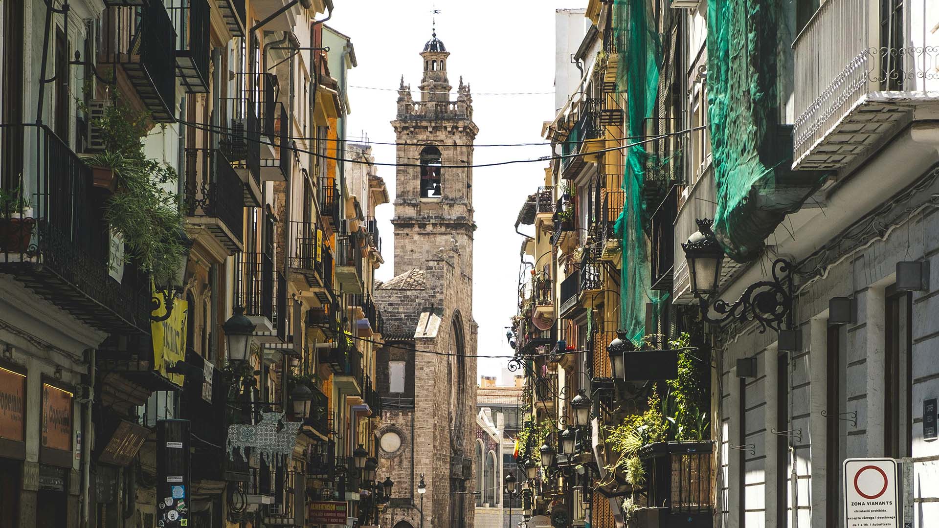 Valencia city street during the day. In the foreground, at the bottom, people are walking in different directions. Shops can be seen on either side. In the background, a church tower stands out against the skyline; it is built of stone, with a bell and a pointed top. Middle class