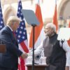 Donald Trump, a la izquierda (vestido con un traje azul, camisa blanca y corbata roja), y Narendra Modi, a la derecha (vestido con un kurta blanco y un chaleco negro), se dan la mano durante una declaración conjunta en un evento oficial al aire libre en Nueva Delhi. Detrás de ellos, varias banderas de los Estados Unidos y la India están colocadas en fila, ondeando ligeramente. En primer plano, hay dos atriles de madera con micrófonos y el emblema nacional de la India grabado en ellos. Modi