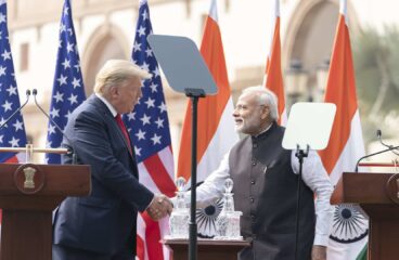 Donald Trump, a la izquierda (vestido con un traje azul, camisa blanca y corbata roja), y Narendra Modi, a la derecha (vestido con un kurta blanco y un chaleco negro), se dan la mano durante una declaración conjunta en un evento oficial al aire libre en Nueva Delhi. Detrás de ellos, varias banderas de los Estados Unidos y la India están colocadas en fila, ondeando ligeramente. En primer plano, hay dos atriles de madera con micrófonos y el emblema nacional de la India grabado en ellos. Modi