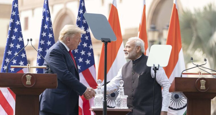 Donald Trump, a la izquierda (vestido con un traje azul, camisa blanca y corbata roja), y Narendra Modi, a la derecha (vestido con un kurta blanco y un chaleco negro), se dan la mano durante una declaración conjunta en un evento oficial al aire libre en Nueva Delhi. Detrás de ellos, varias banderas de los Estados Unidos y la India están colocadas en fila, ondeando ligeramente. En primer plano, hay dos atriles de madera con micrófonos y el emblema nacional de la India grabado en ellos. Modi