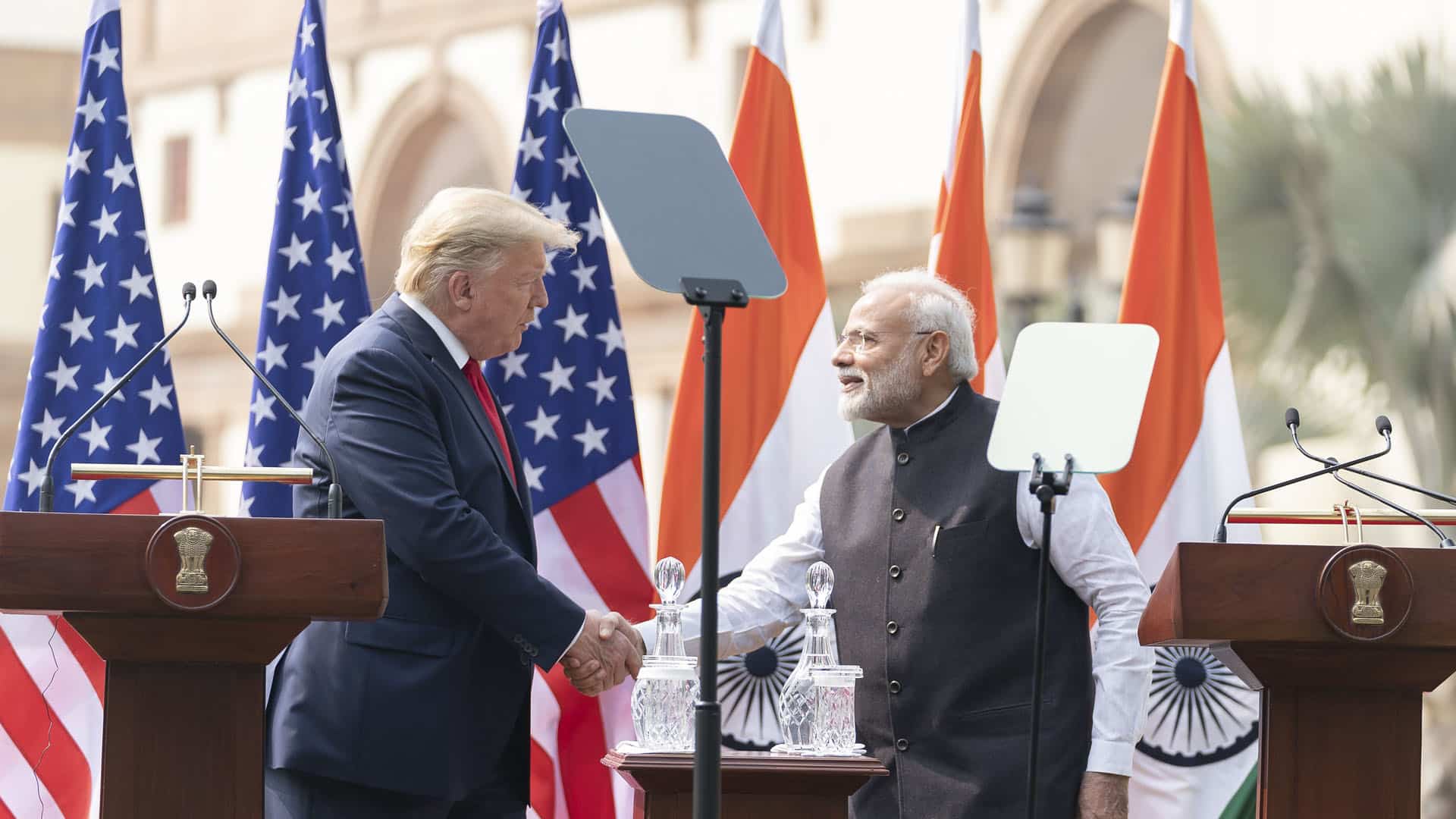 Donald Trump, a la izquierda (vestido con un traje azul, camisa blanca y corbata roja), y Narendra Modi, a la derecha (vestido con un kurta blanco y un chaleco negro), se dan la mano durante una declaración conjunta en un evento oficial al aire libre en Nueva Delhi. Detrás de ellos, varias banderas de los Estados Unidos y la India están colocadas en fila, ondeando ligeramente. En primer plano, hay dos atriles de madera con micrófonos y el emblema nacional de la India grabado en ellos. Modi