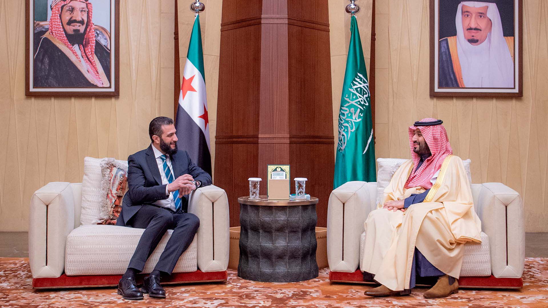 El príncipe heredero saudí Mohammed bin Salman (derecha) se reúne con el presidente interino de Siria, Ahmed al-Sharaa (izquierda), en Riad el 2 de febrero de 2025. Ambos están sentados en sillones blancos con banderas de Siria y Arabia Saudí detrás y retratos de líderes saudíes en la pared. Siria