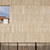 Fachada de un edificio de hormigón gris con textura vertical y una gran ventana cuadrada abierta que enmarca un edificio de ladrillos al fondo con un balcón cerrado de ventanas blancas. En la parte inferior de la fachada, una reja de lamas verticales de color negro