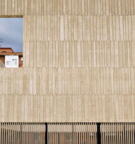 Fachada de un edificio de hormigón gris con textura vertical y una gran ventana cuadrada abierta que enmarca un edificio de ladrillos al fondo con un balcón cerrado de ventanas blancas. En la parte inferior de la fachada, una reja de lamas verticales de color negro