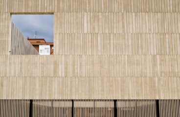 Fachada de un edificio de hormigón gris con textura vertical y una gran ventana cuadrada abierta que enmarca un edificio de ladrillos al fondo con un balcón cerrado de ventanas blancas. En la parte inferior de la fachada, una reja de lamas verticales de color negro