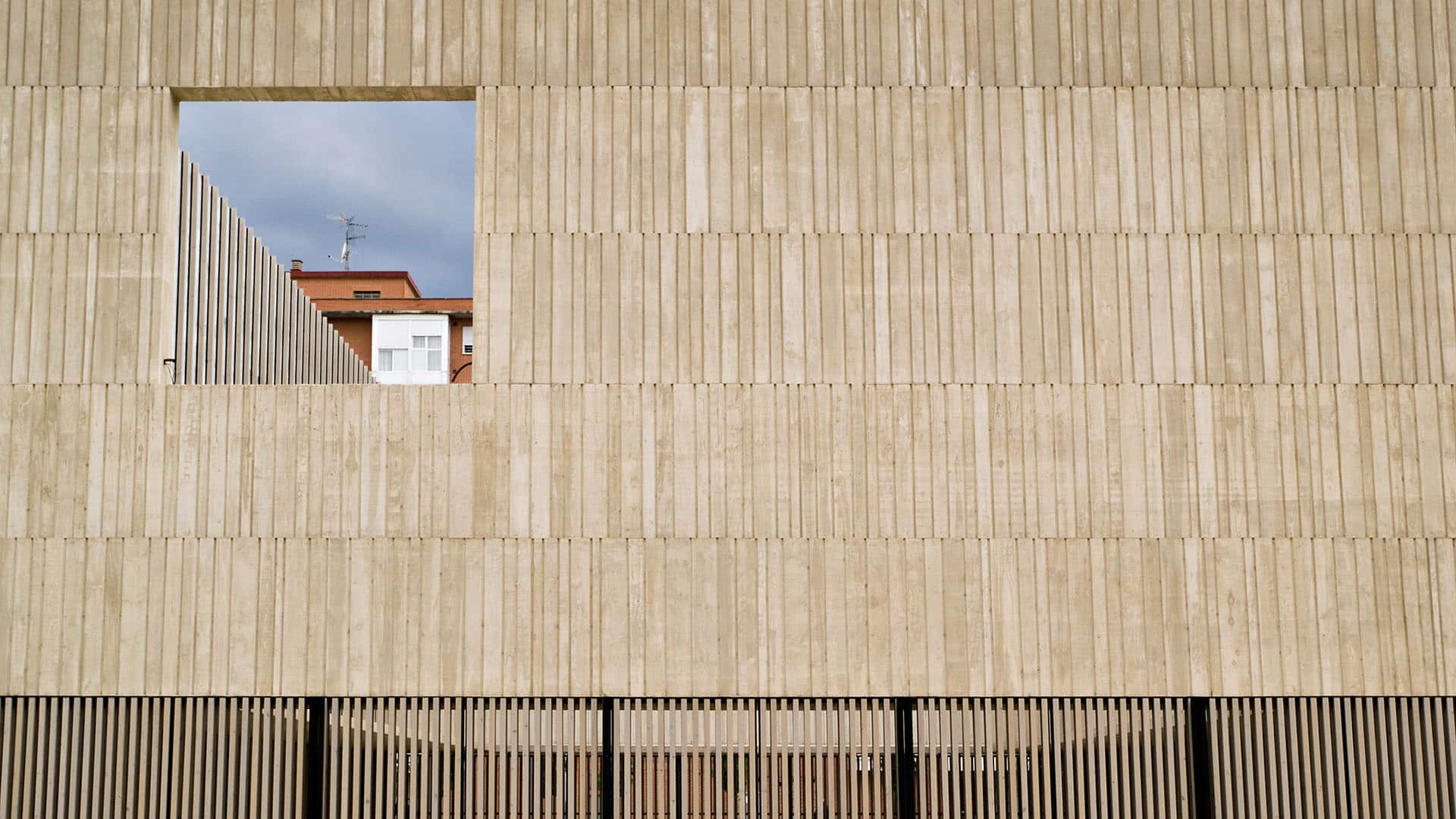 Fachada de un edificio de hormigón gris con textura vertical y una gran ventana cuadrada abierta que enmarca un edificio de ladrillos al fondo con un balcón cerrado de ventanas blancas. En la parte inferior de la fachada, una reja de lamas verticales de color negro