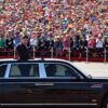 En primer plano, Xi Jinping (de pie, vestido con un traje oscuro) saluda desde un vehículo negro con detalles plateados en las ventanillas y una bandera china ondeando en la parte delantera, durante el desfile militar en las celebraciones del Día de la Victoria en China, el 3 de septiembre de 2015. En el fondo, una multitud ondea banderas y aplaude mientras el presidente participa en el evento oficial en un día soleado. China