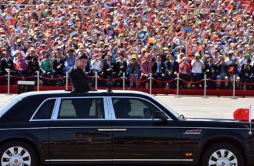 En primer plano, Xi Jinping (de pie, vestido con un traje oscuro) saluda desde un vehículo negro con detalles plateados en las ventanillas y una bandera china ondeando en la parte delantera, durante el desfile militar en las celebraciones del Día de la Victoria en China, el 3 de septiembre de 2015. En el fondo, una multitud ondea banderas y aplaude mientras el presidente participa en el evento oficial en un día soleado. China