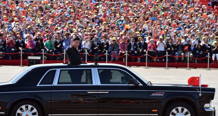 En primer plano, Xi Jinping (de pie, vestido con un traje oscuro) saluda desde un vehículo negro con detalles plateados en las ventanillas y una bandera china ondeando en la parte delantera, durante el desfile militar en las celebraciones del Día de la Victoria en China, el 3 de septiembre de 2015. En el fondo, una multitud ondea banderas y aplaude mientras el presidente participa en el evento oficial en un día soleado. China