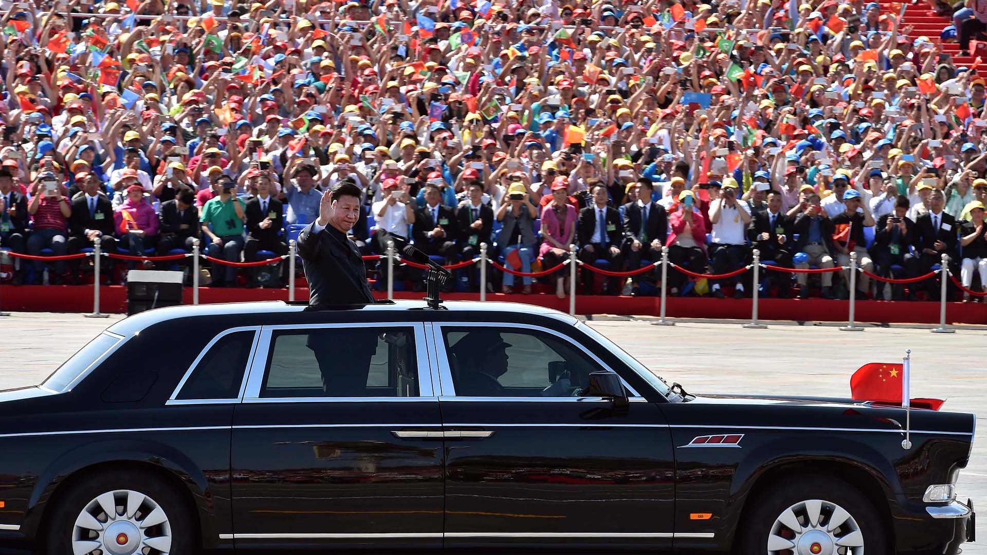 En primer plano, Xi Jinping (de pie, vestido con un traje oscuro) saluda desde un vehículo negro con detalles plateados en las ventanillas y una bandera china ondeando en la parte delantera, durante el desfile militar en las celebraciones del Día de la Victoria en China, el 3 de septiembre de 2015. En el fondo, una multitud ondea banderas y aplaude mientras el presidente participa en el evento oficial en un día soleado. China