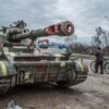 Tanque militar de diferentes tonalidades de verde, aparcado en la cuneta de una carretera en la ciudad ucraniana de Chernígov. En el lateral derecho se aprecia a un hombre con chaqueta oscura, pantalones y gorro negros, caminando con una bicicleta cerca del tanque. En el fondo, se ve un coche de color cobre y un viandante, bajo un cielo nublado. Ucrania