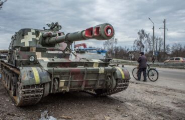 Tanque militar de diferentes tonalidades de verde, aparcado en la cuneta de una carretera en la ciudad ucraniana de Chernígov. En el lateral derecho se aprecia a un hombre con chaqueta oscura, pantalones y gorro negros, caminando con una bicicleta cerca del tanque. En el fondo, se ve un coche de color cobre y un viandante, bajo un cielo nublado. Ucrania