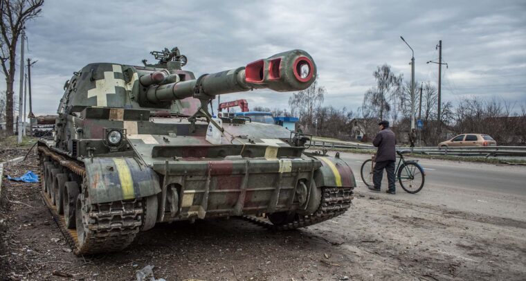 Tanque militar de diferentes tonalidades de verde, aparcado en la cuneta de una carretera en la ciudad ucraniana de Chernígov. En el lateral derecho se aprecia a un hombre con chaqueta oscura, pantalones y gorro negros, caminando con una bicicleta cerca del tanque. En el fondo, se ve un coche de color cobre y un viandante, bajo un cielo nublado. Ucrania