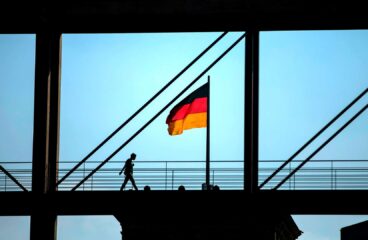 Silueta de una persona caminando sobre una pasarela enmarcada por una estructura que aparece de color negro. La bandera nacional de Alemania (tricolor con tres franjas: negra, roja y amarilla) sobre el techo del Reichstag en Berlín, bajo un cielo soleado