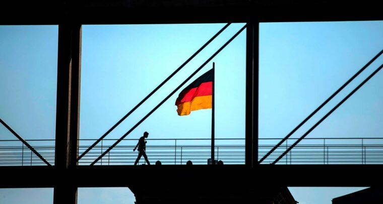 Silueta de una persona caminando sobre una pasarela enmarcada por una estructura que aparece de color negro. La bandera nacional de Alemania (tricolor con tres franjas: negra, roja y amarilla) sobre el techo del Reichstag en Berlín, bajo un cielo soleado