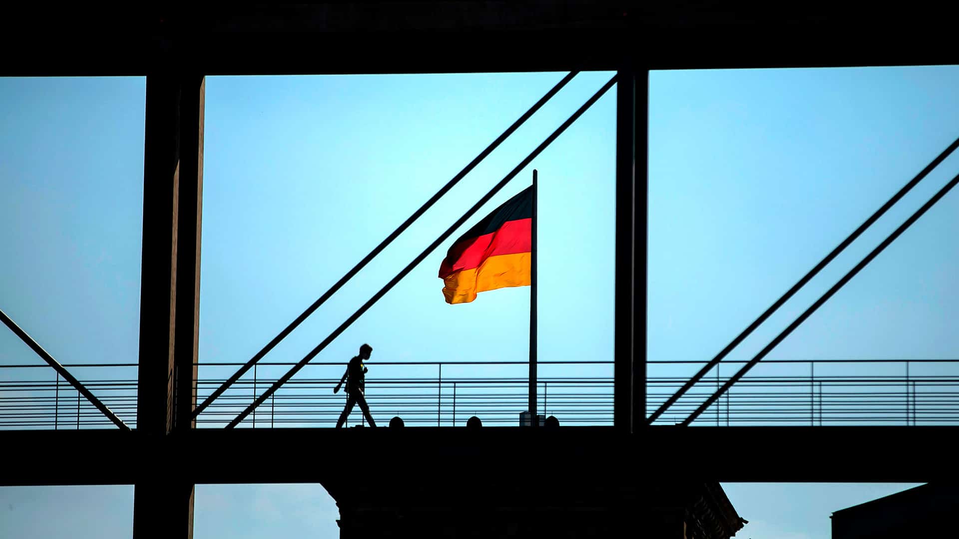 Silueta de una persona caminando sobre una pasarela enmarcada por una estructura que aparece de color negro. La bandera nacional de Alemania (tricolor con tres franjas: negra, roja y amarilla) sobre el techo del Reichstag en Berlín, bajo un cielo soleado